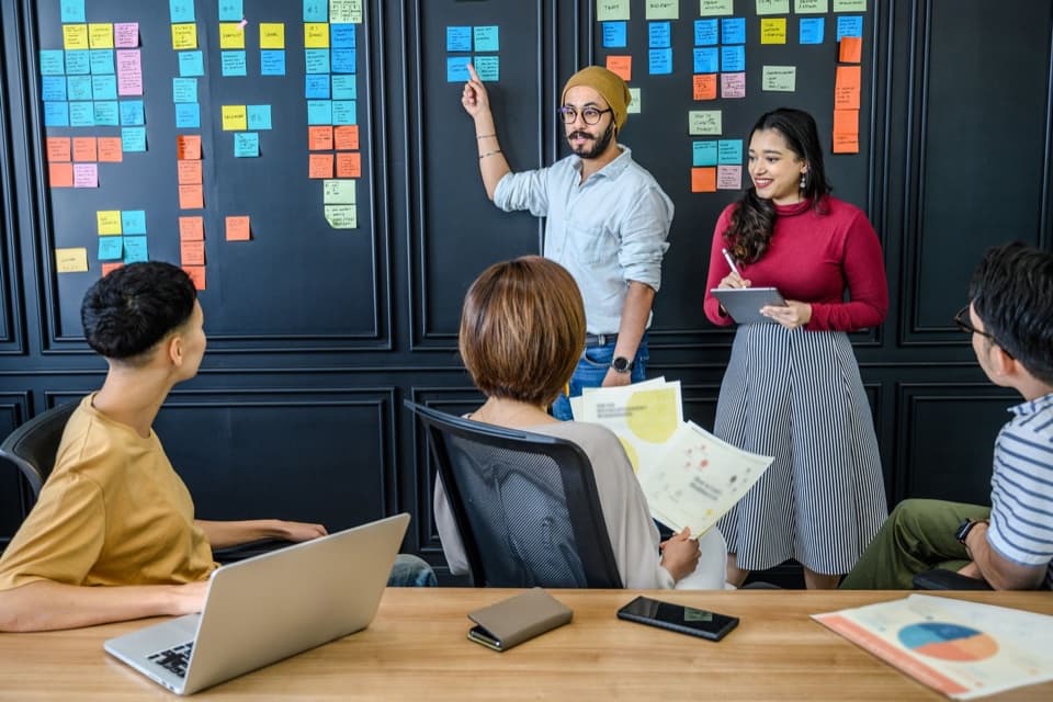 Homem aponta um mural de post-its que mapeia um processo, ao lado de colega com tablet, diante do grupo
