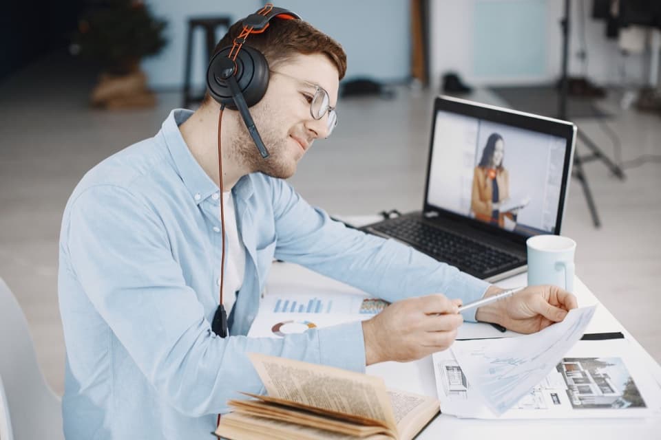 Homem de headset anota com caneta enquanto assiste a uma aula em vídeo no laptop, com livro aberto